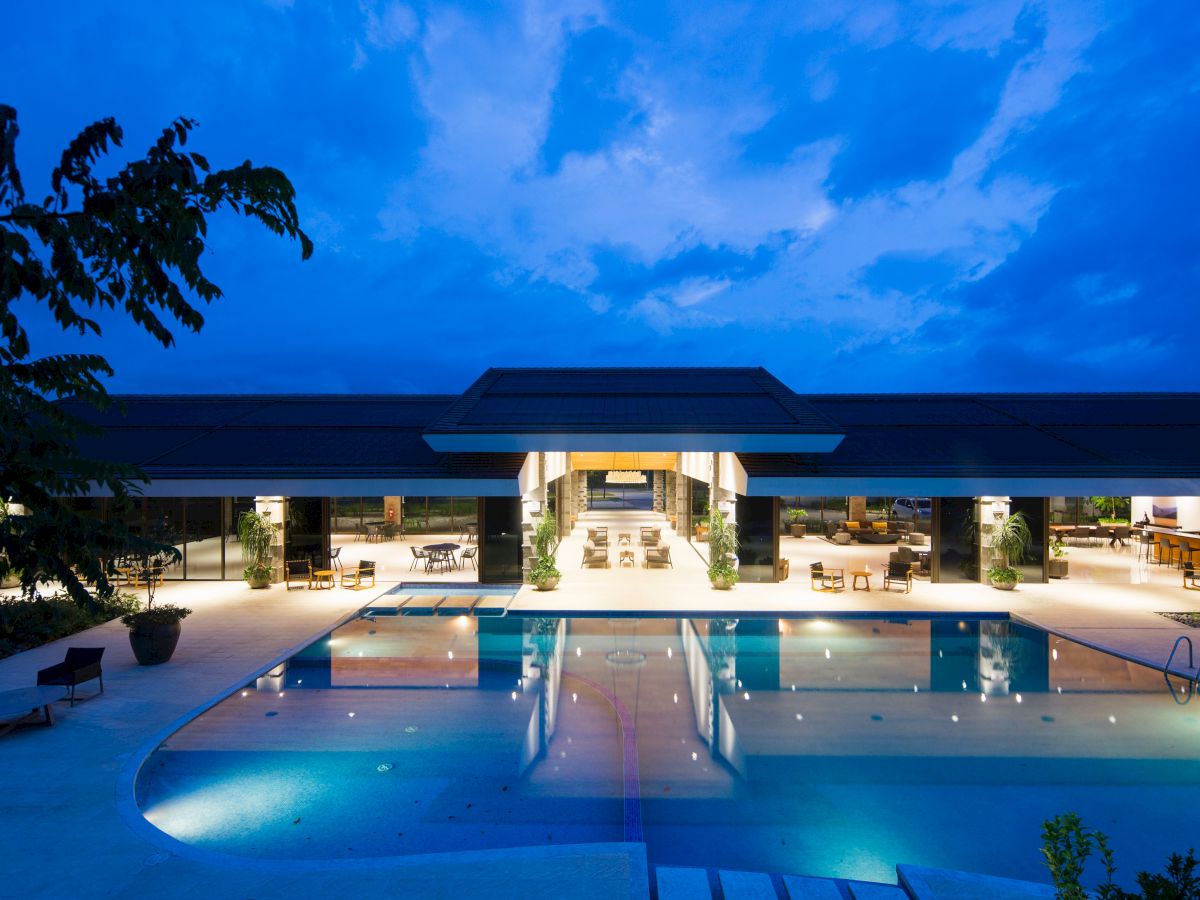 A brightly lit modern building is shown at dusk with a large, inviting swimming pool in the foreground, surrounded by lounge chairs.
