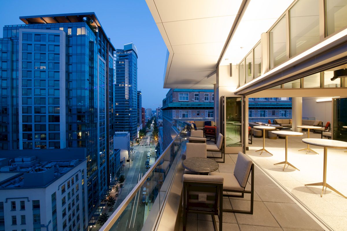 A modern cityscape view at twilight from a balcony with chairs, tables, and buildings nearby.
