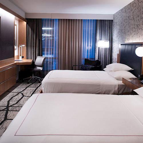 A modern hotel room with two neatly made beds, a flat-screen TV, a work desk, and large windows with curtains, creating a comfortable and stylish space.