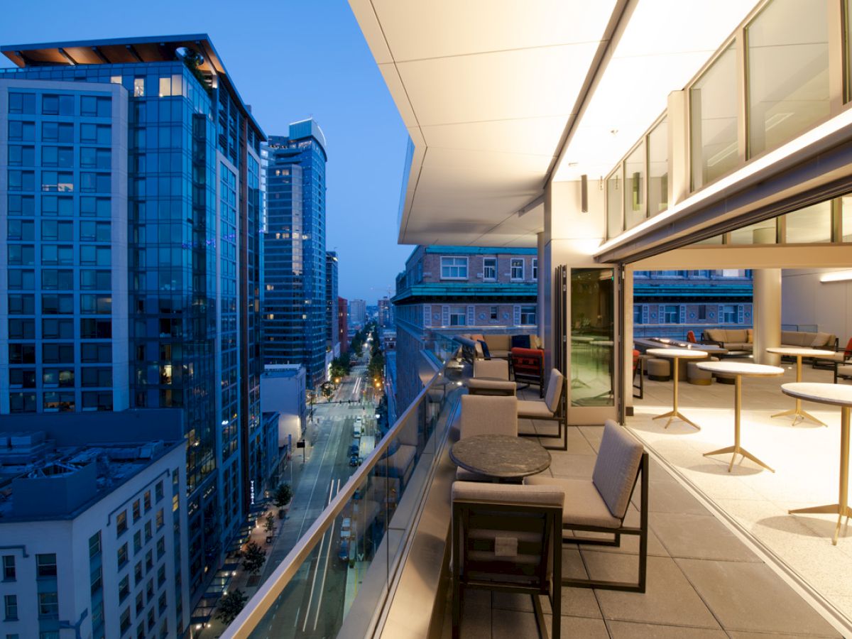 A modern balcony with outdoor seating overlooks a city street lined with tall buildings at dusk. The interior space is visible, well-lit and furnished.