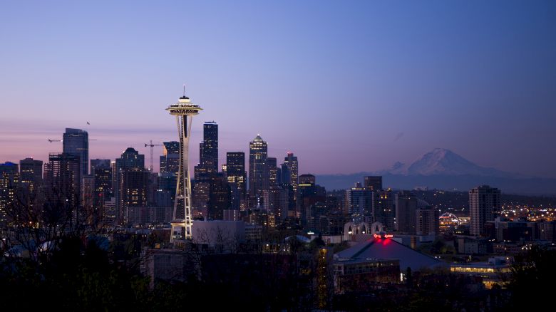 The image shows a cityscape of Seattle at dusk, featuring the Space Needle with Mount Rainier in the background.