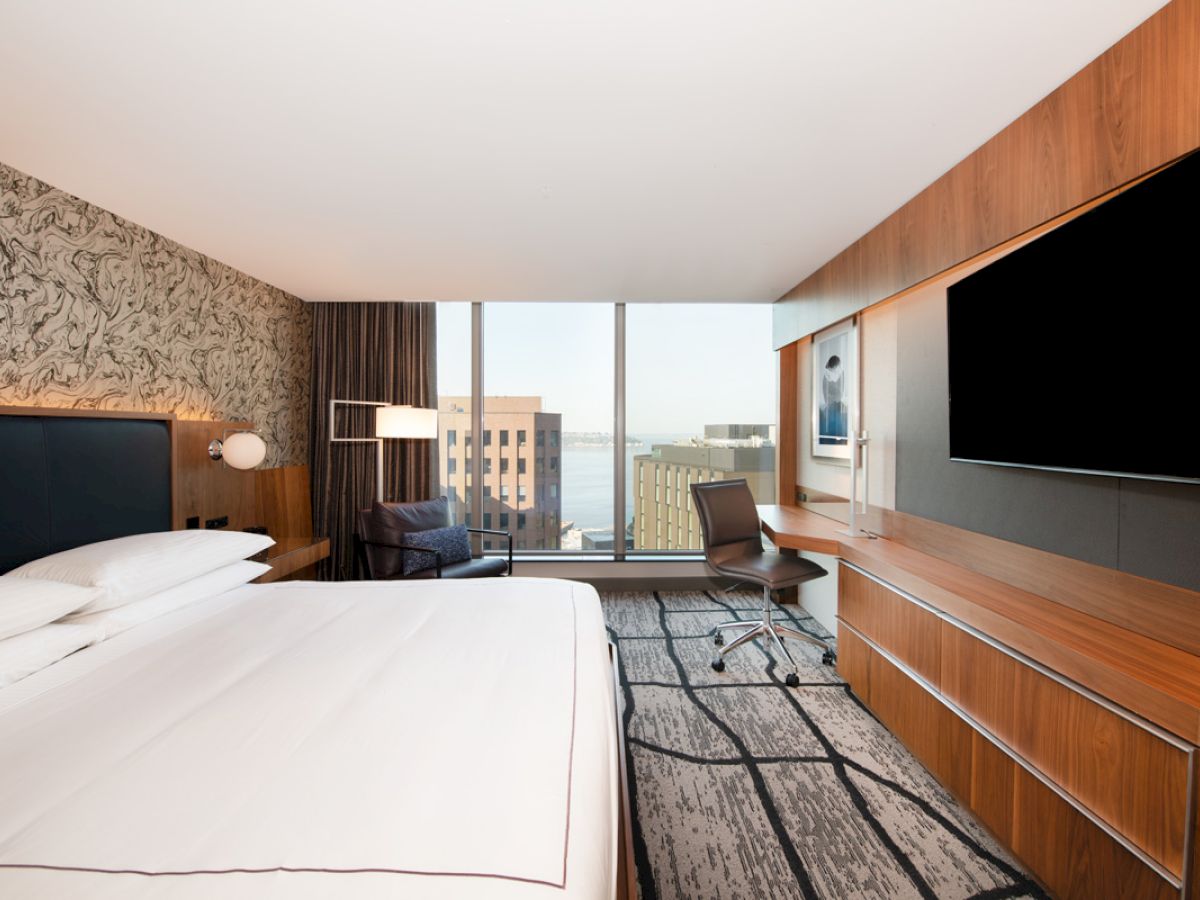 A modern hotel room with a large bed, desk, chair, and flat-screen TV. A window offers a view of buildings and a body of water in the background.