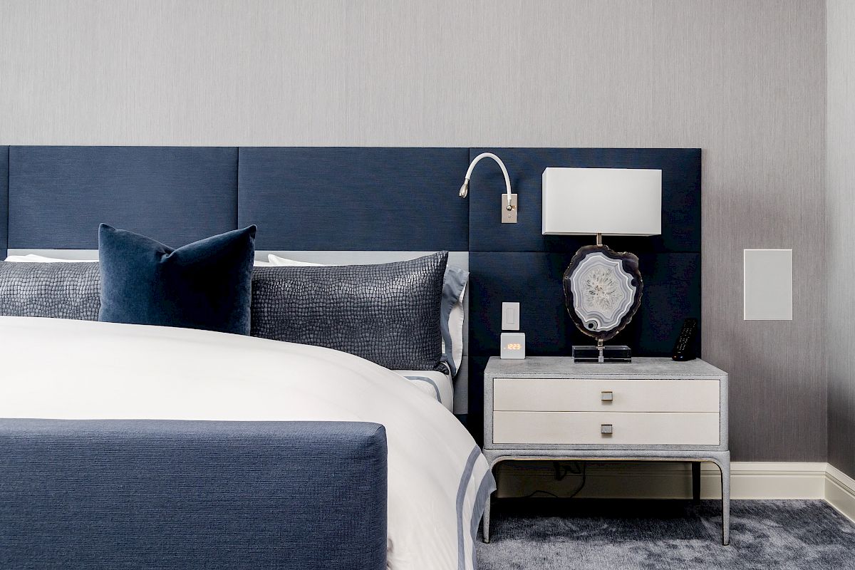 A modern bedroom featuring a blue upholstered headboard, white bedding, and a nightstand with a lamp and decorative item.