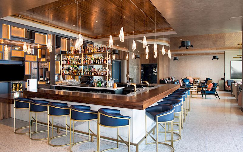 A modern bar with wooden accents, elegant lighting, a stocked liquor shelf, and blue bar stools. Cozy seating area in the background, ending the sentence.