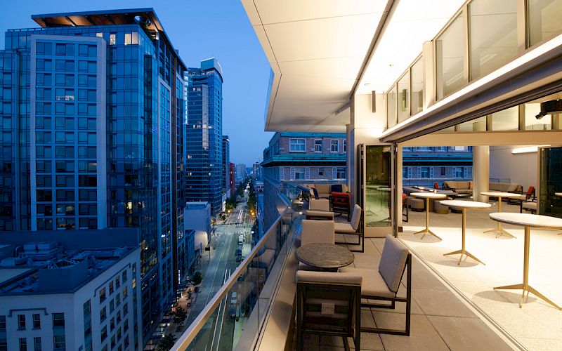 A modern cityscape at dusk, with a balcony featuring outdoor seating and tables, overlooking a street lined with tall buildings and lit windows.
