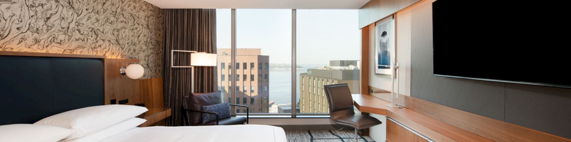 A modern hotel room with a large bed, a wall-mounted TV, a desk and chair, and large windows offering a city and water view.
